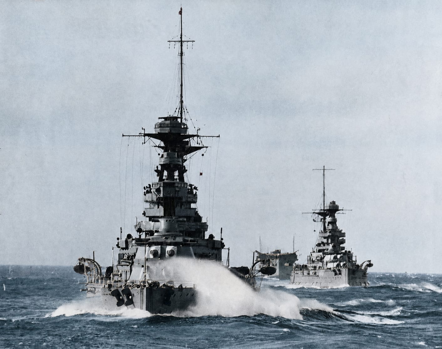 HMS Barham driving through a swell with HMS Malaya astern, and the flush-decked aircraft carrier HMS Argus on the horizon between them
